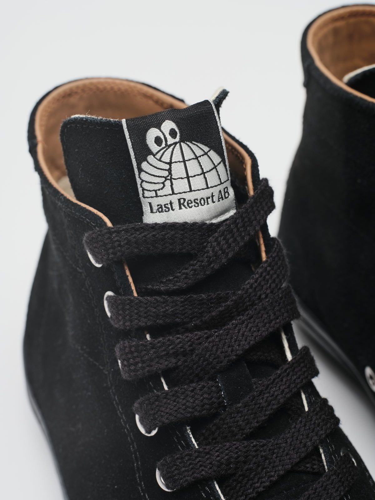 Close-up of a black suede high-top sneaker with black laces and a white tongue label featuring a cartoon face and the text 'Last Resort 48'.