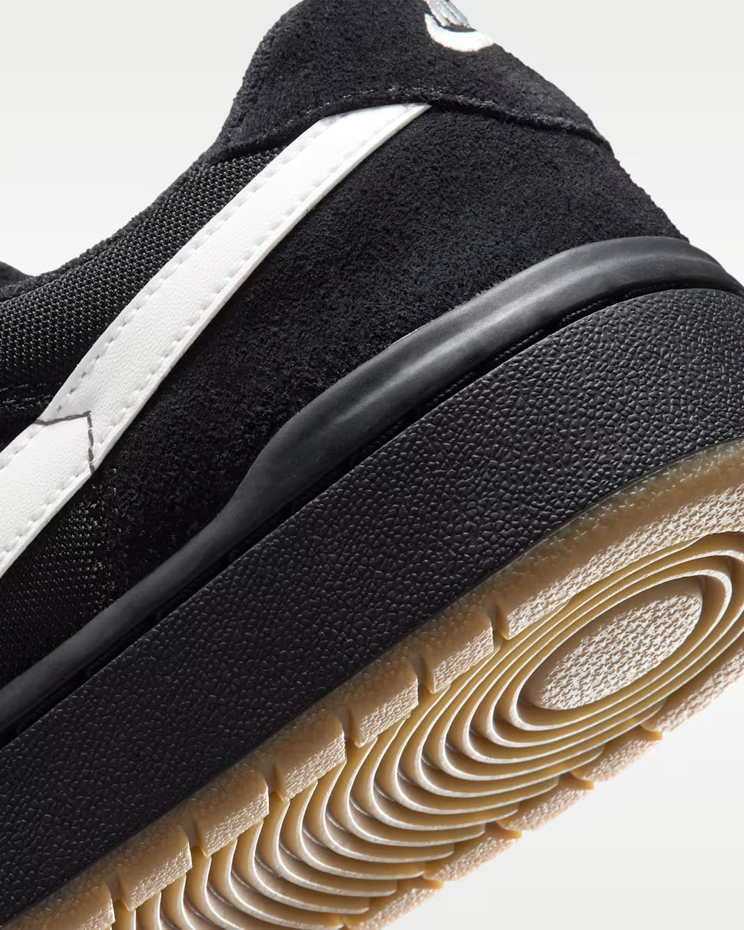 Close-up of a black suede and mesh sneaker with a white side stripe and a tan gum outsole, photographed on a light gray background.