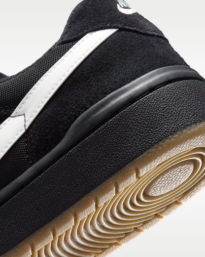 Close-up of a black suede and mesh sneaker with a white side stripe and a tan gum outsole, photographed on a light gray background.