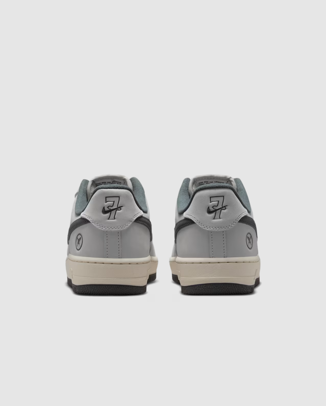 A pair of low-top sneakers shown from the back, featuring a light gray leather upper with dark gray overlays, a cream midsole, and a black outsole, set against a light gray background.