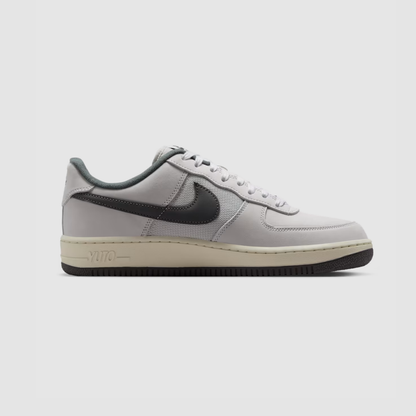 A Nike Air Force 1 Low sneaker in light gray and black, featuring a light gray leather upper, black Swoosh, and a cream-colored midsole with black outsole.